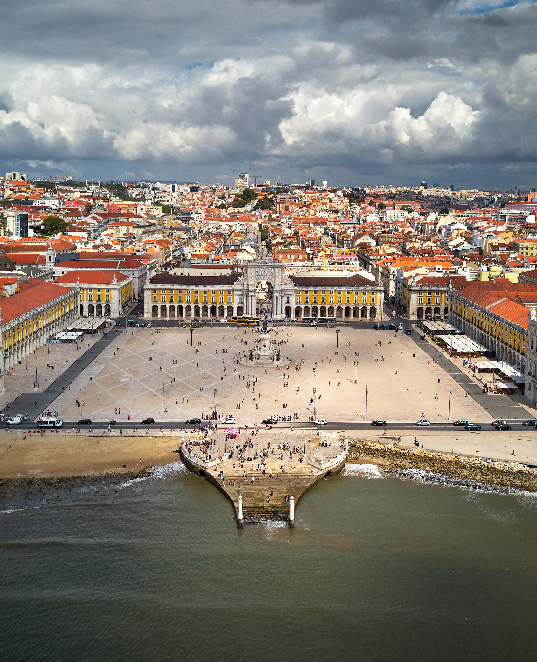 En esta imagen vemos a todo color una ciudad costera justo al lado de una playa de la que sobresale un dique peatonal. Observamos una gran plaza cuadrada rodeada de edificios neoclásicos. Vemos una urbe salpicada de colores rojos teja y blancos de muros y fachadas. Está cruzada por doquier por una red de calles pequeñas y paseables. Al fondo el cielo se muestra gris debido a algunas nubes de textura algodonosa. Además  grupos de turistas pasean por doquier.