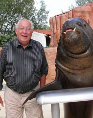 Un hombre mayor  de aspecto alegre y rechoncho nos observa de pie mientras posa junto a una gran foca oscura. El hombre viste una camisa negra a rayas y esta algo arremangado. Abulta su gran vientre y destaca su sonriente rostro. Tiene el pelo cano y gafas. Abre su boca como si estuviera comentando anécdotas alegres. Él y el mamífero marino se hallan al aire libre  en un espacio exterior  aunque al fondo se aprecian casas y muros rurales.