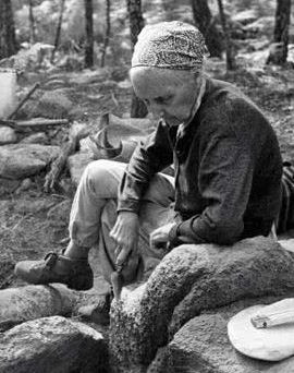 En la imagen aparece una mujer anciana y delgada trabajando al aire libre en una zona arbolada de suelo de piedra y musgo. Viste confortable con pantalón y jersey de sport. Lleva un pañuelo en su cabeza protegiendo su pelo. Calza unas robustas botas de alpinismo y lleva en su mano una suerte de espátula con la que hurga entre las piedras a las que dedica toda su atención y concentración.