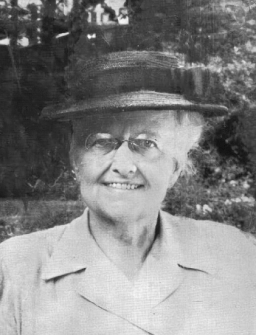 Una anciana mujer posa con un sombrero a principios del siglo XX. Se trata de una foto oscura y manchada  pero que se ilumina con su ligera sonrisa y con su clara blusa de aspecto de exploradora. Tiene gafas redondas  pelo blanco que se alborota bajo su sombrero  y barbilla levemente afilada. Se nos muestra con porte recto y amable en un entorno abierto y natural.